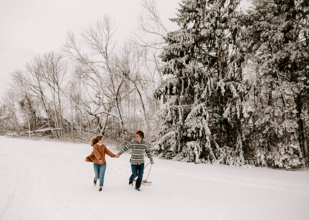 Iowa Winter Snowy Engagement Session, Couple runs through the snow with a sled