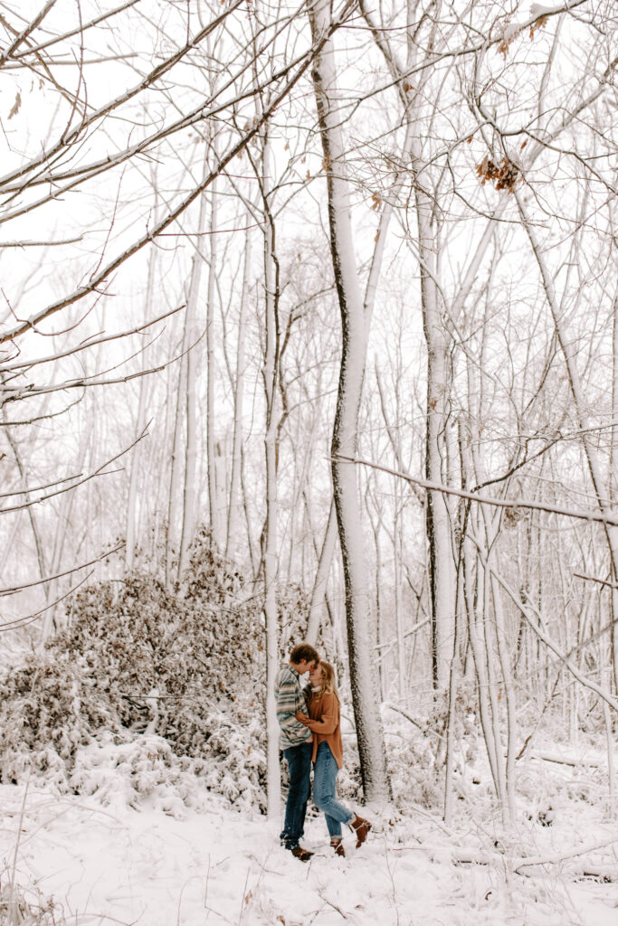 Couple goes in for a kiss in the snow covered woods, Iowa winter engagement photos