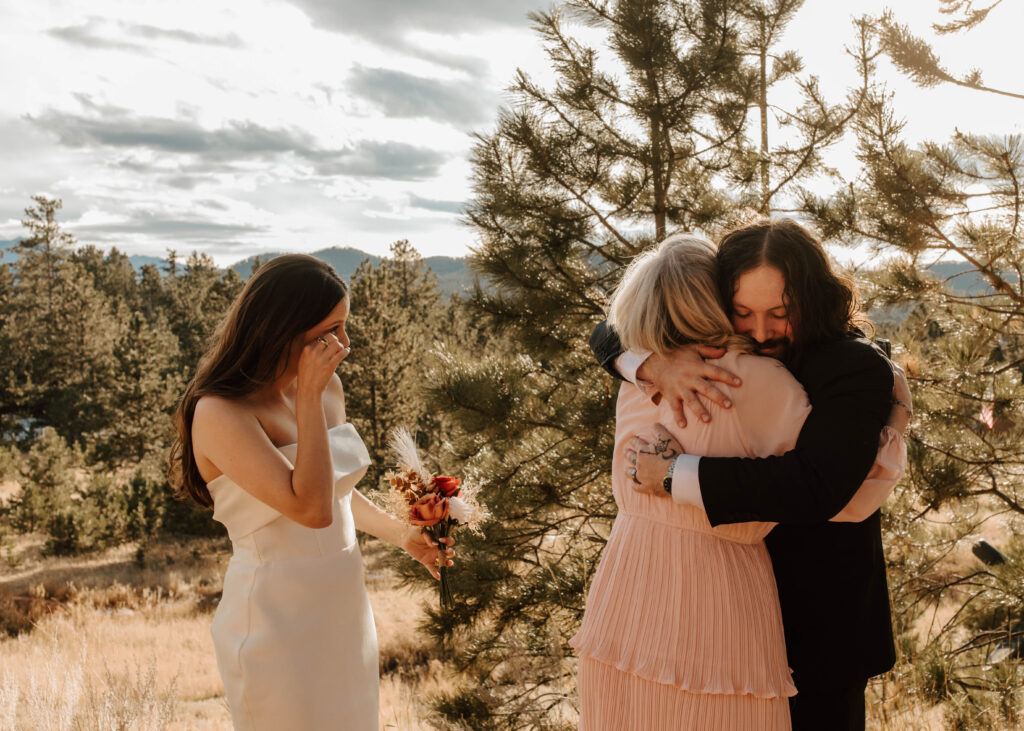 groom and mother hug while bride cries happy tears with mountains behind them

