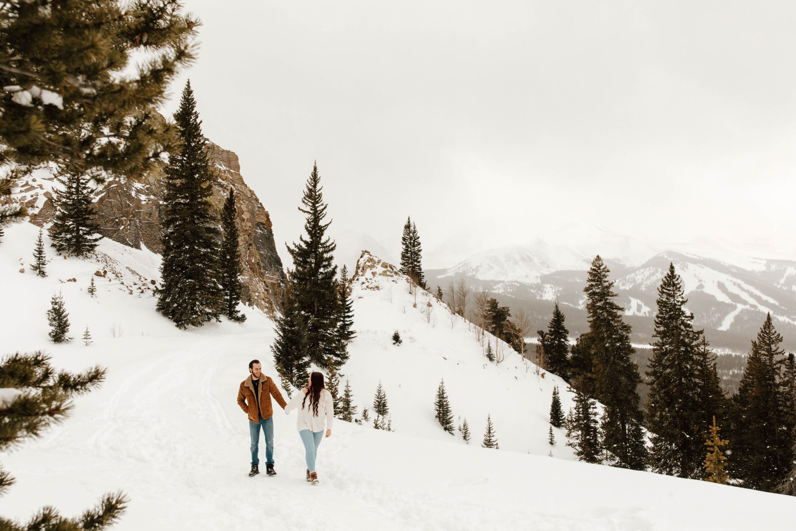 couple walks through the snow in Breckenridge, she leads him. the mountains are in the distance