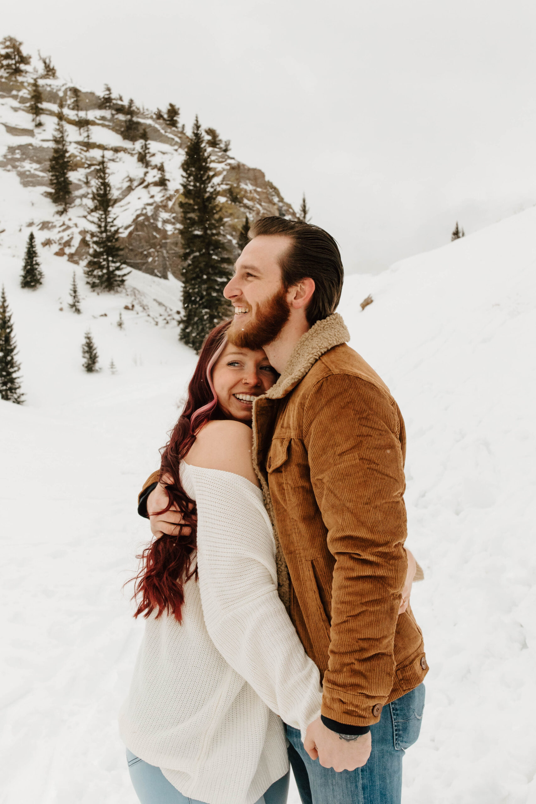 couple embraces, they both are laughing. there is snow falling over them and the mountains