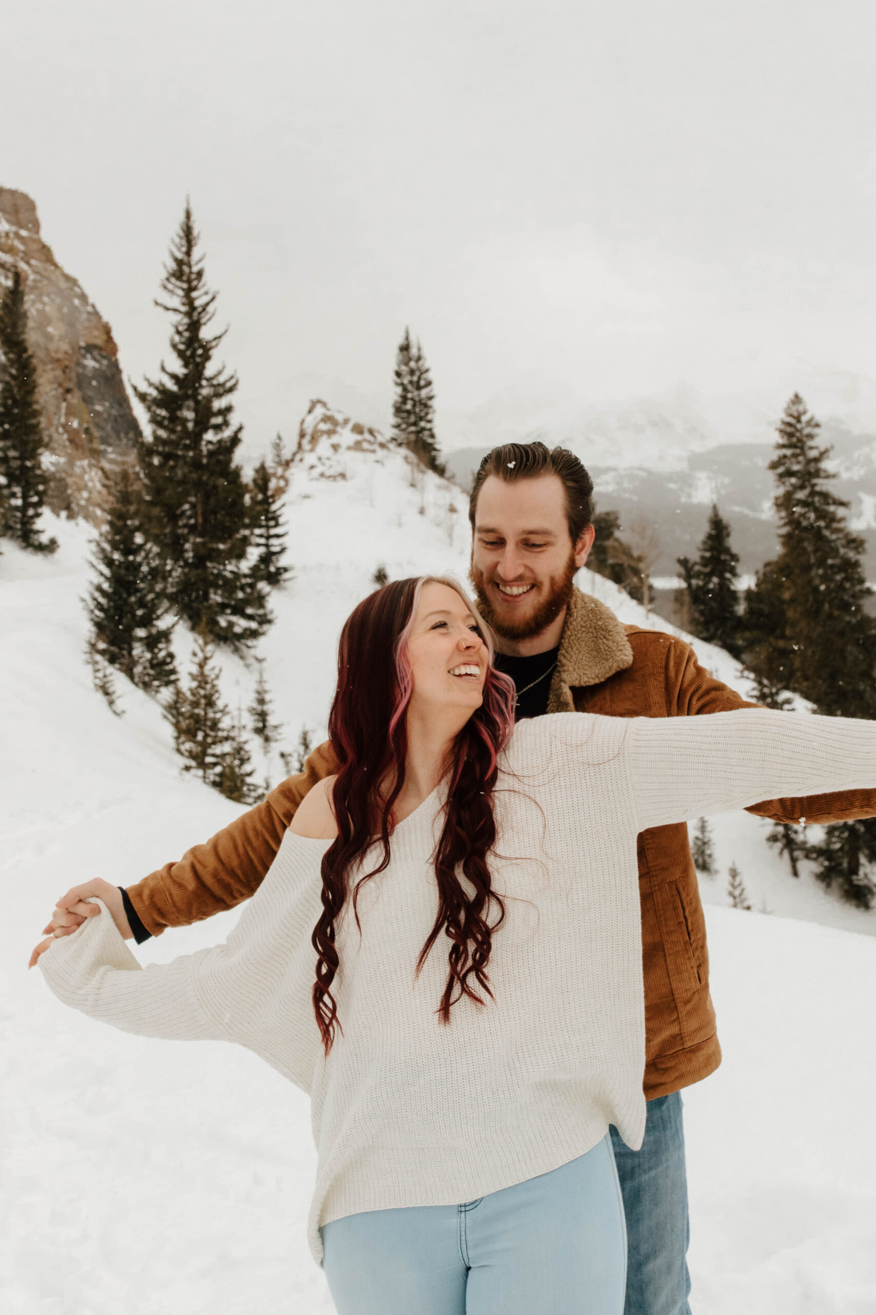 couple holds hands with their arms out, they are both laughing and are in the mountains