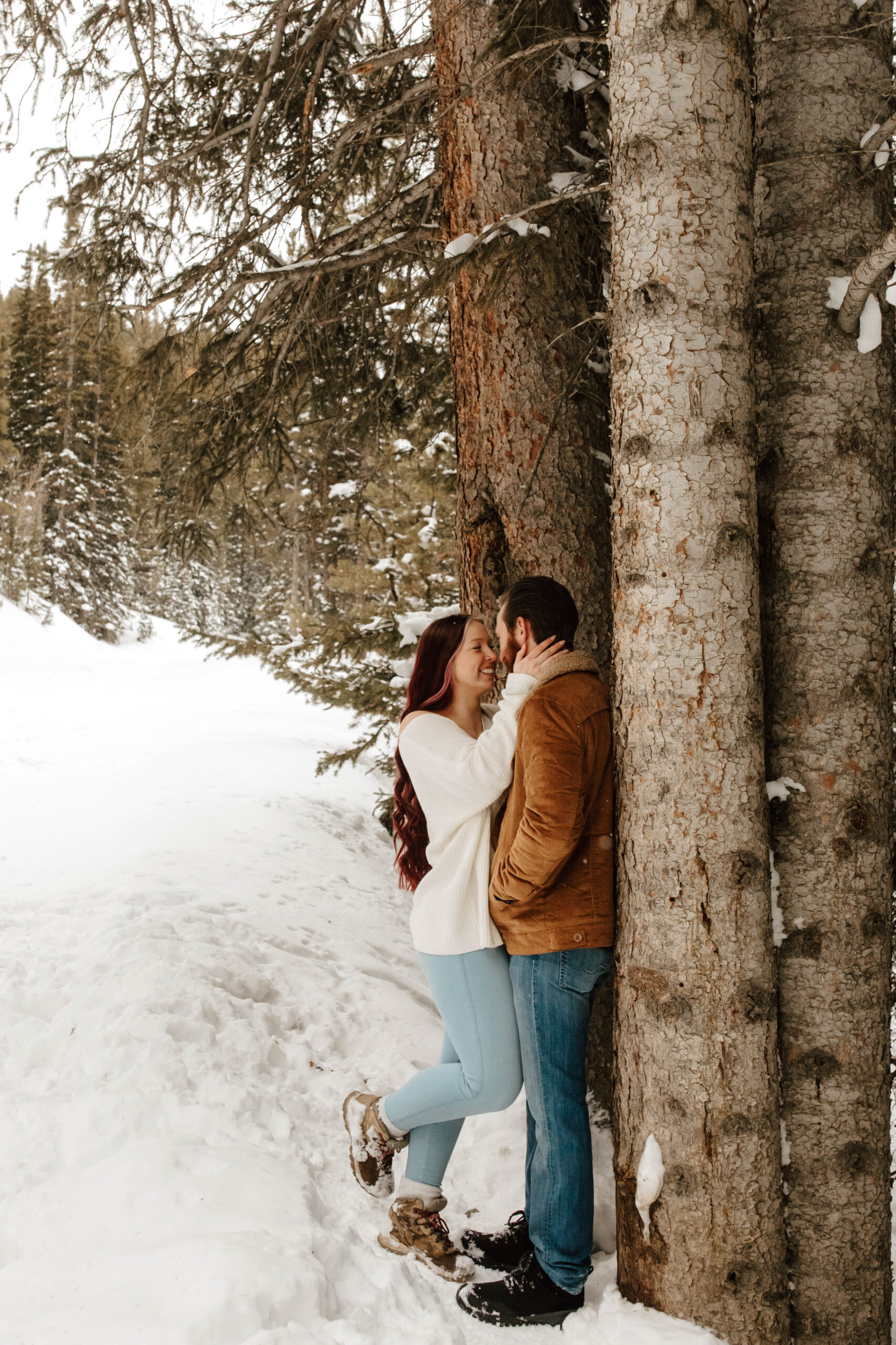 couple snuggles up together against a tree, surrounded by snow. her leg is popped