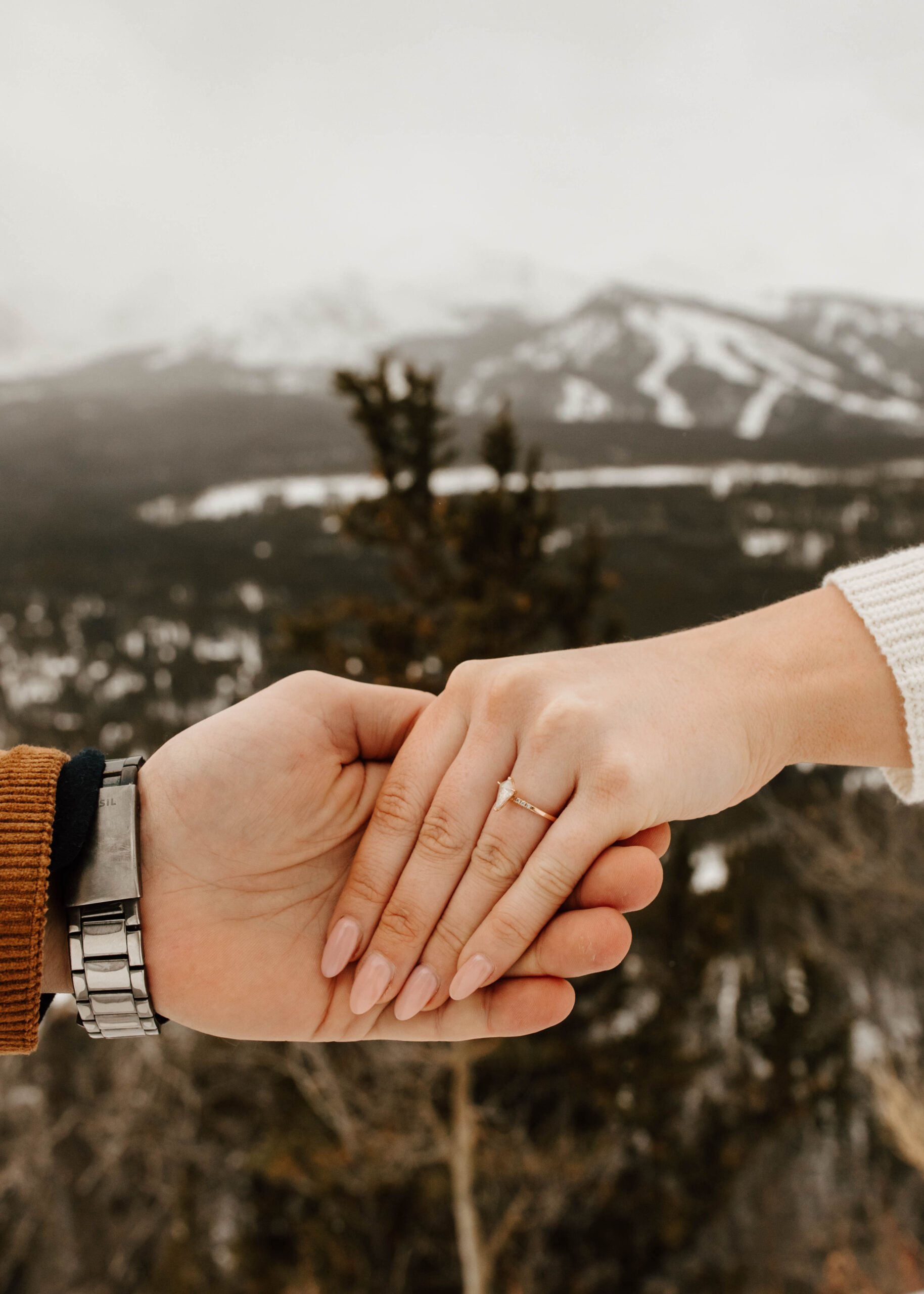 the couple is holding hands, her engagement ring is the star of the show in front of the snowy mountain backdrop