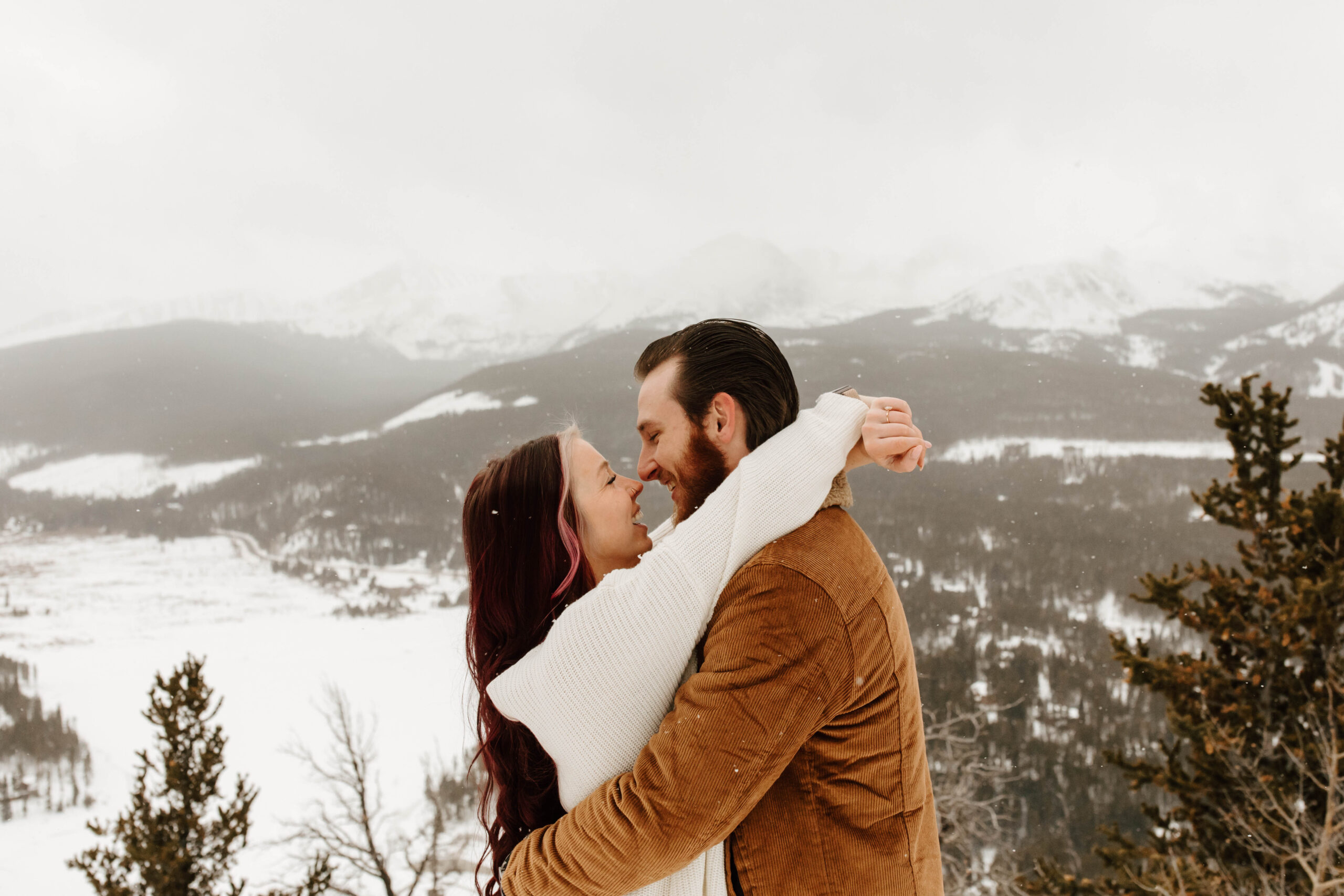 couple hugs and smiles at each other with a look of love, while the snow falls over them and the mountains in the distance