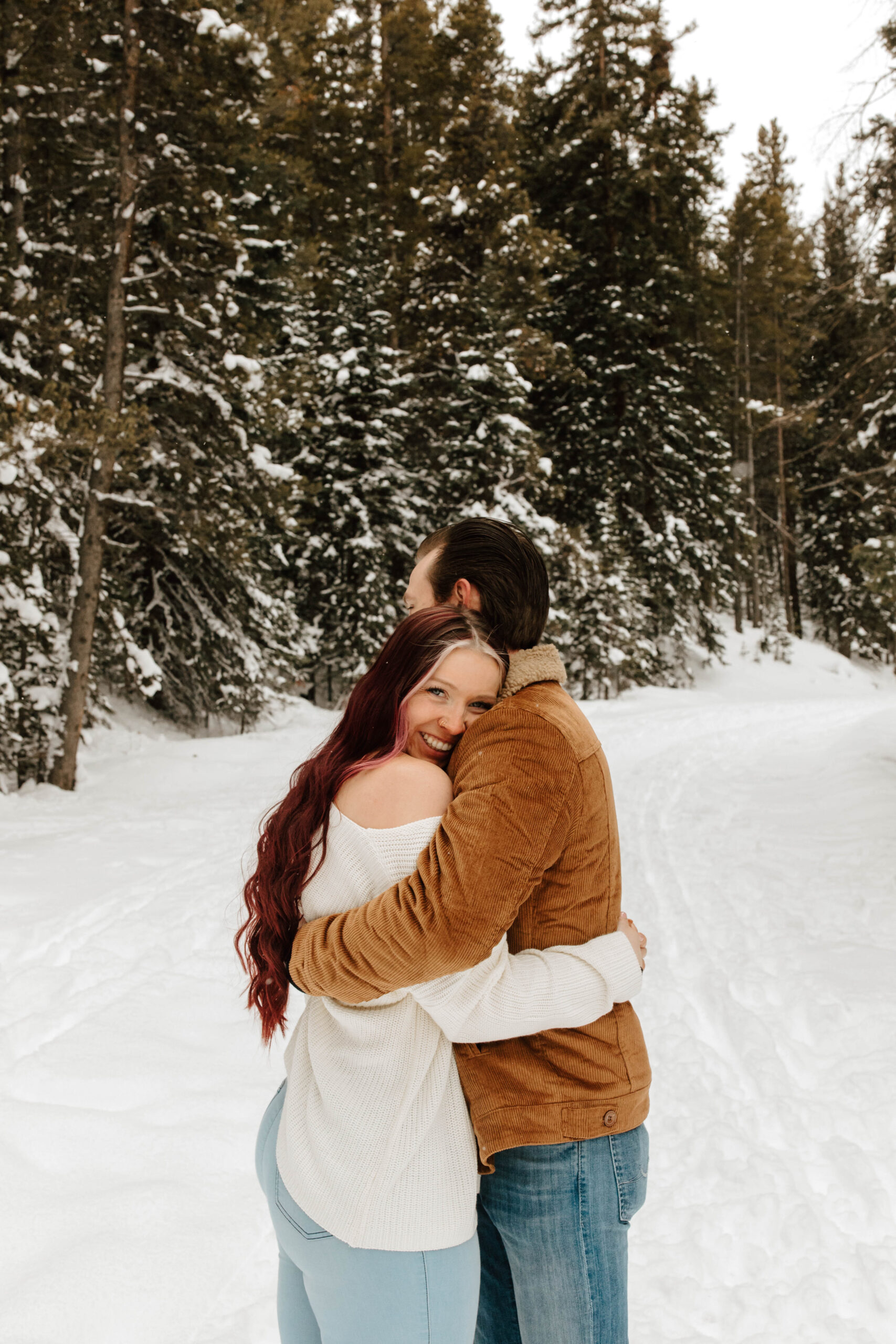 couple hugs, and she smiles at the camera. they are surrounded by snow and tall evergreen trees.
