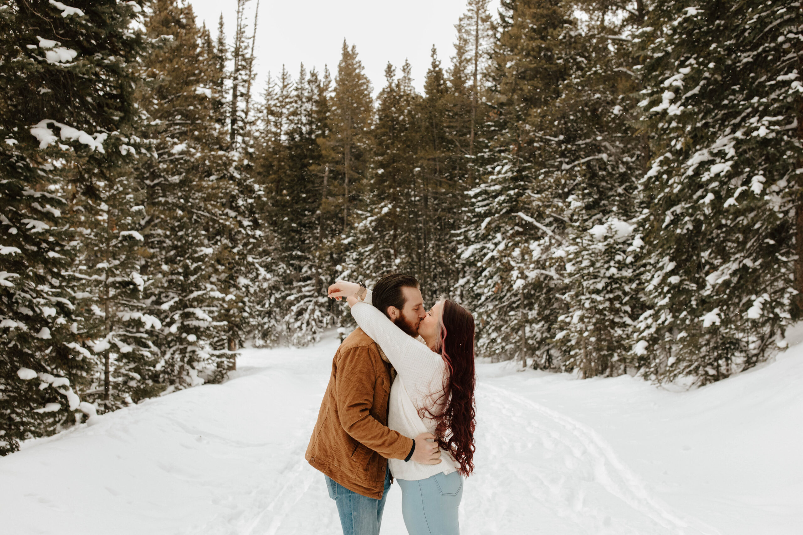 couple kisses while the snow falls around them and the mountains. she has her arms wrapped around his neck