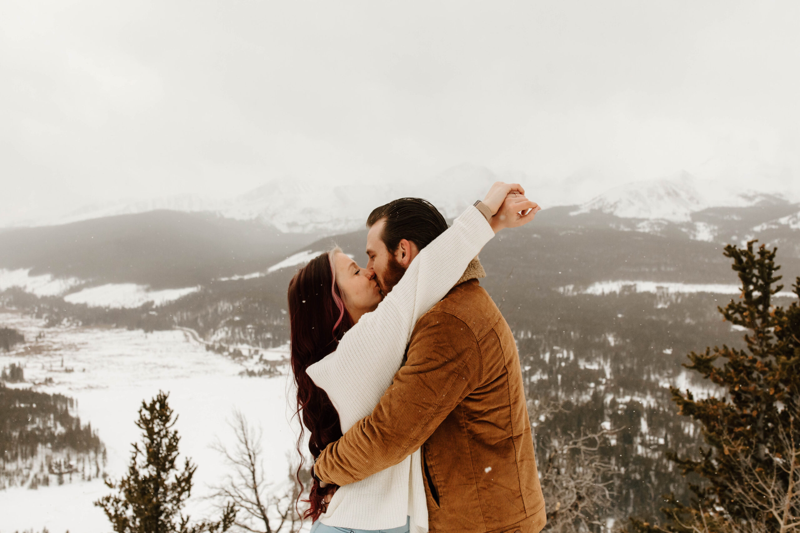 couple kisses with mountains in the background. he wears a brown jacket and she wears a white sweater