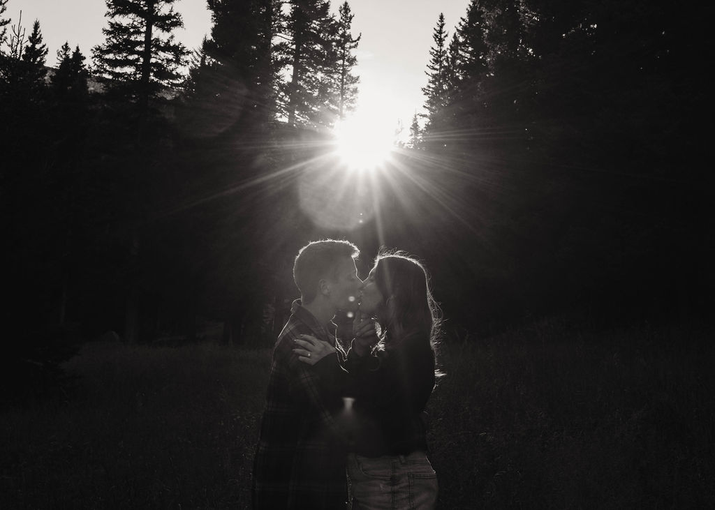 couple kisses as light streams between the trees during engagement photos