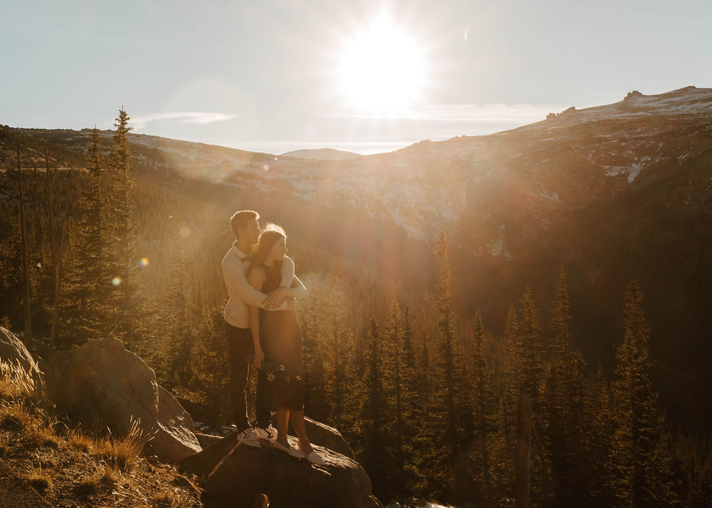 couple stands together hugging on top of mountain during golden hour