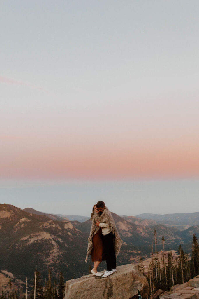 couple snuggles in blanket on top of mountain as the sky is pink