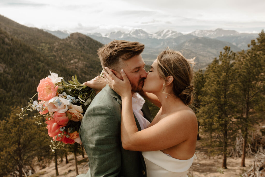 couple kisses with wildflower bouquet and mountains behind them