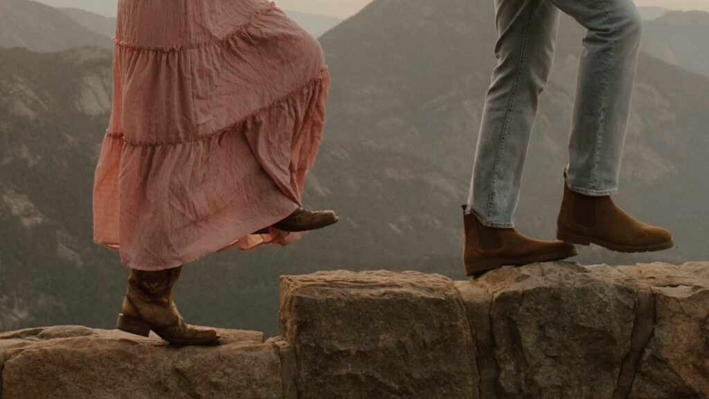 girl in cowgirl boots and a pink skirt walks with her fiance in front of mountains