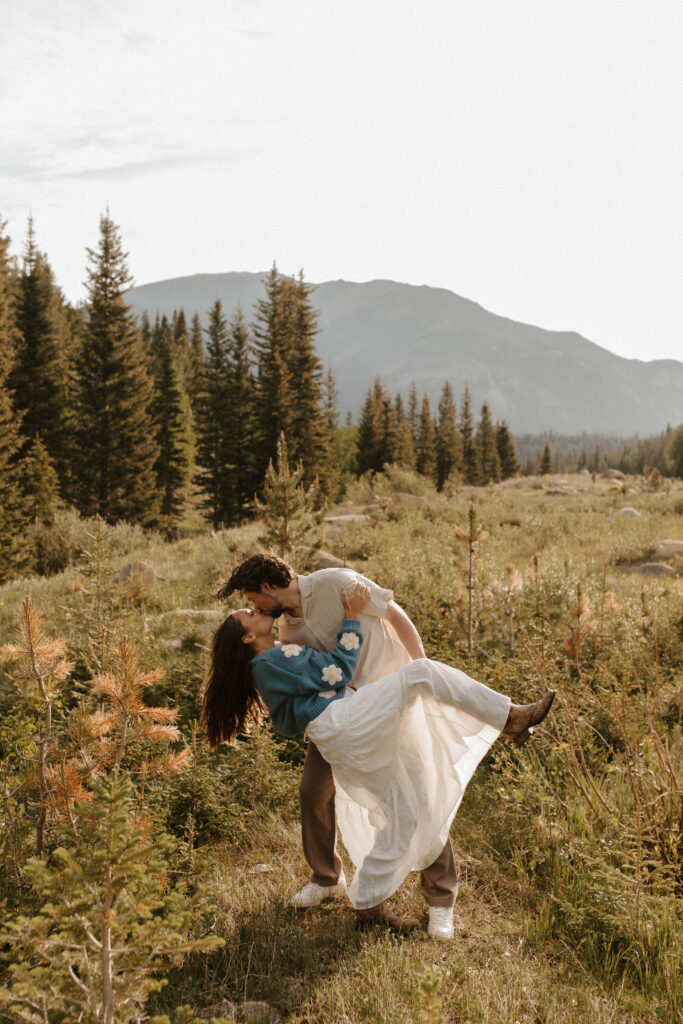 couple dips and kisses during engagement photos in mountain meadow